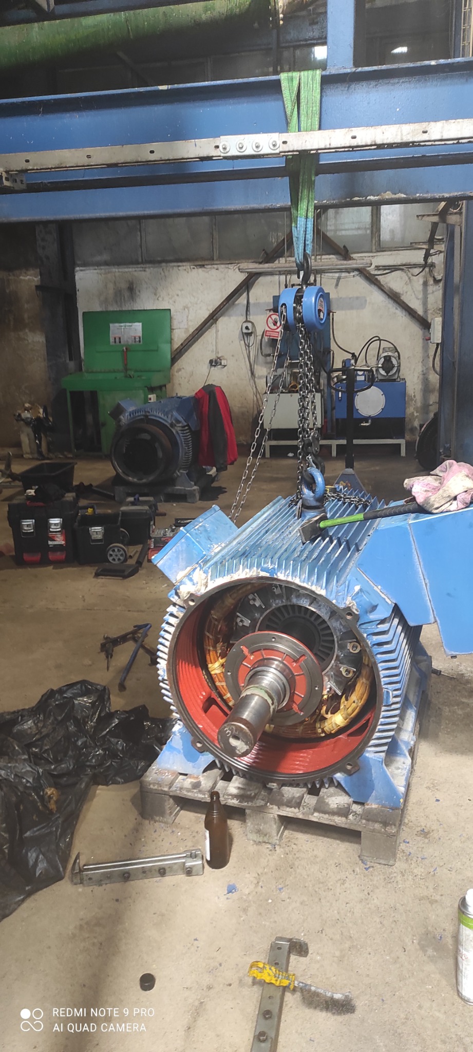 Rozłożony, niebieski silnik elektryczny z czerwonym wnętrzem, podwieszony na suwnicy w warsztacie, widoczne narzędzia i butelka.