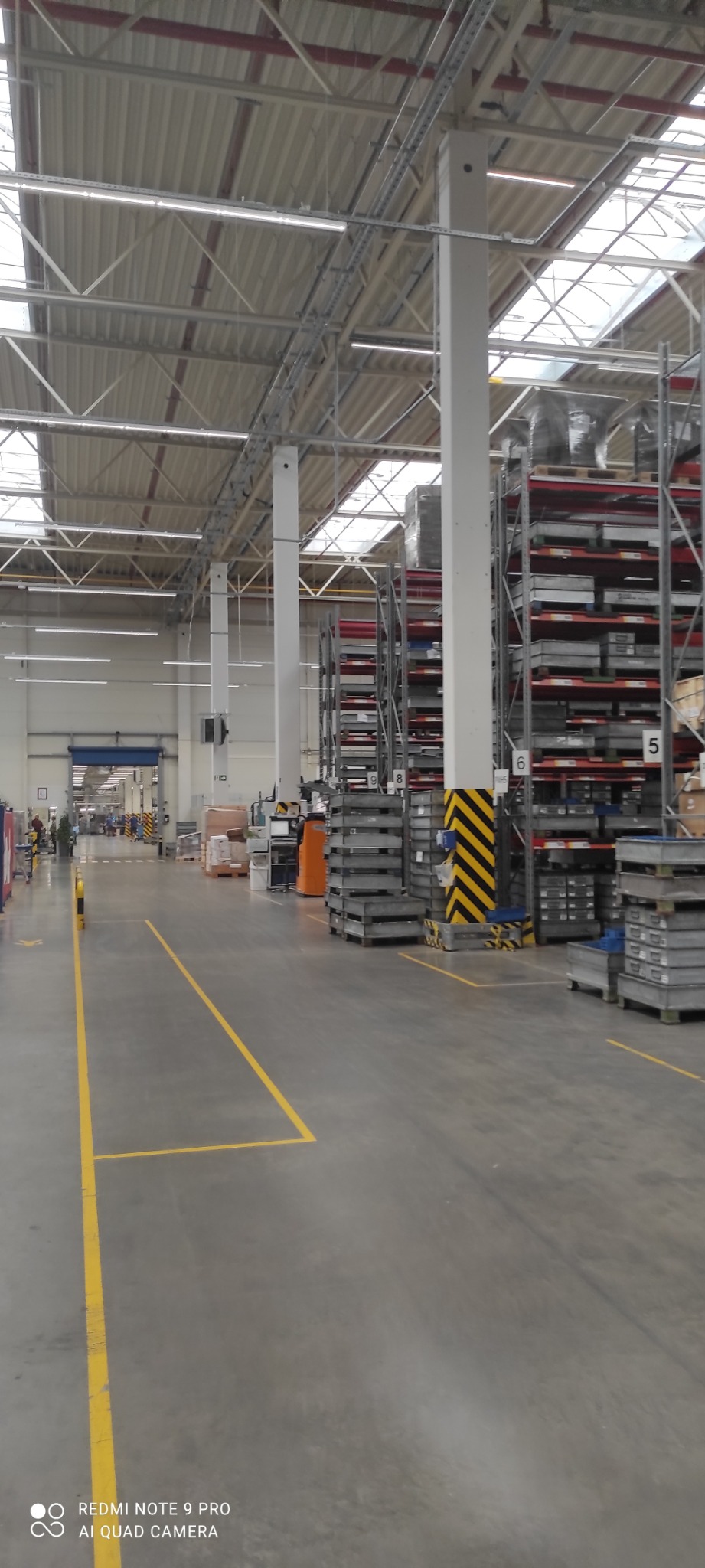 Wnętrze hali magazynowej z regałami wypełnionymi metalowymi elementami, widoczne żółte linie wyznaczające ciągi komunikacyjne i słupy z czarno-żółtymi pasami ostrzegawczymi.