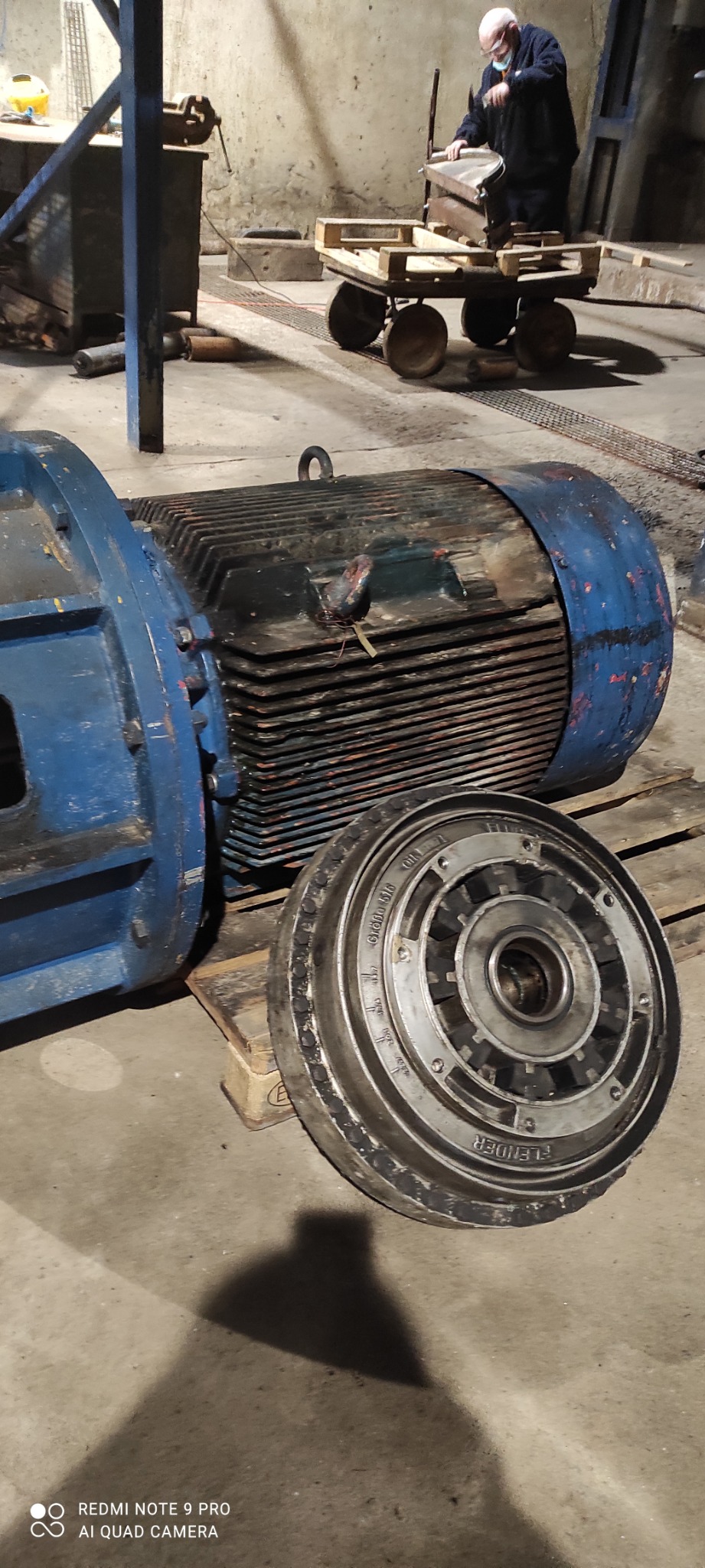 Duży, niebiesko-czarny silnik elektryczny i jego metalowa część (Flender) leżące na drewnianej palecie w warsztacie, w tle majster w masce ochronnej przy pracy.