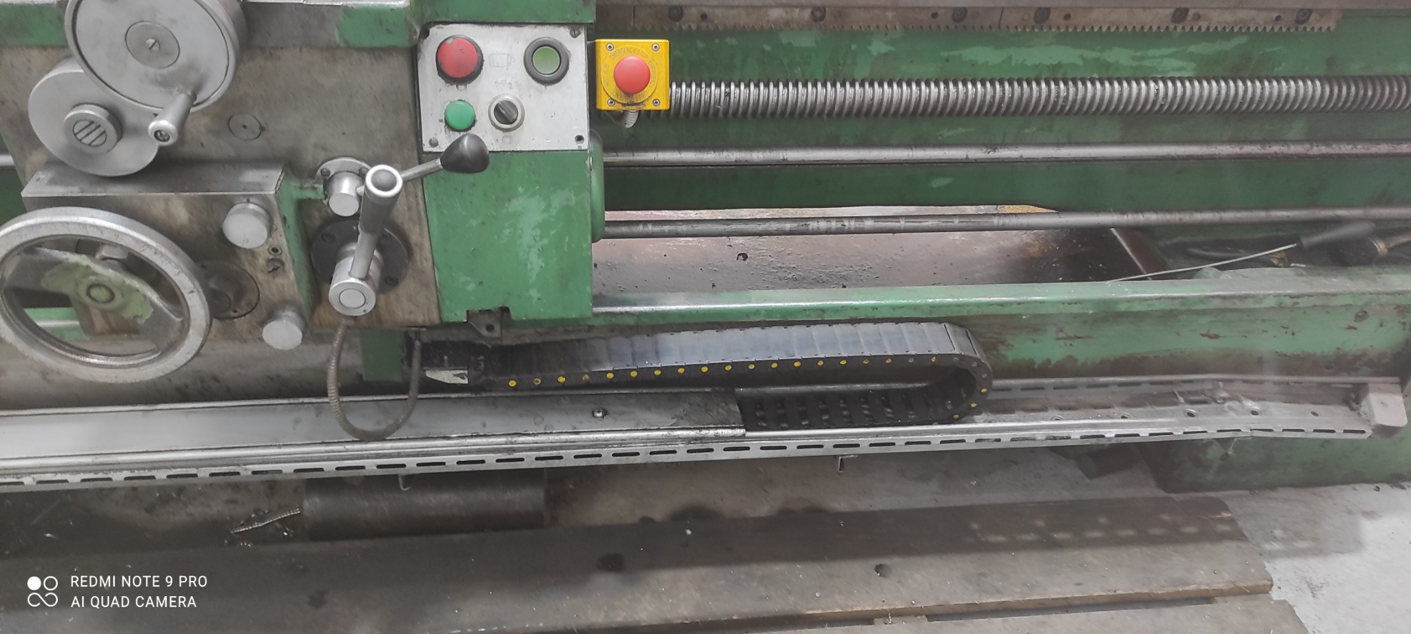 Fragment starej, zielonej tokarki z widocznymi elementami sterującymi, w tym przyciskami awaryjnymi i pokrętłami.