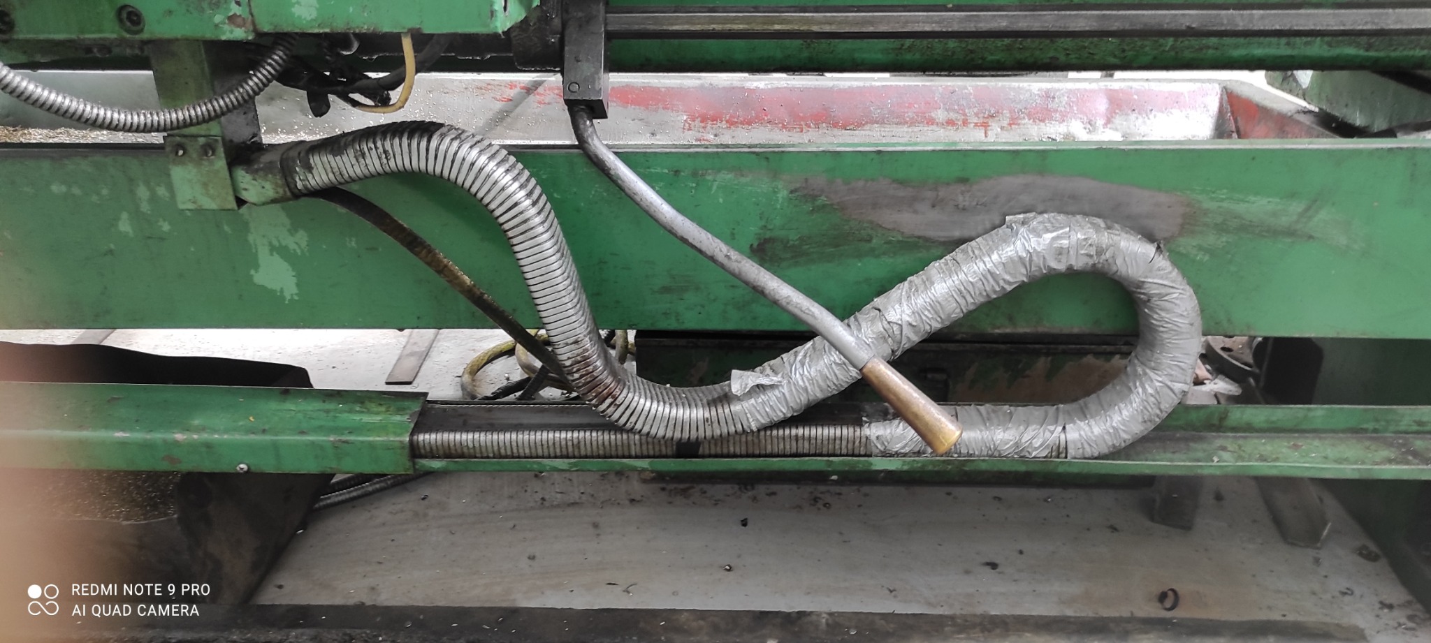 Fragment maszyny przemysłowej z widocznymi przewodami ochronnymi (jeden owinięty taśmą) i metalowym prętem z brązową rękojeścią, na tle zielonej, pomalowanej ramy maszyny.