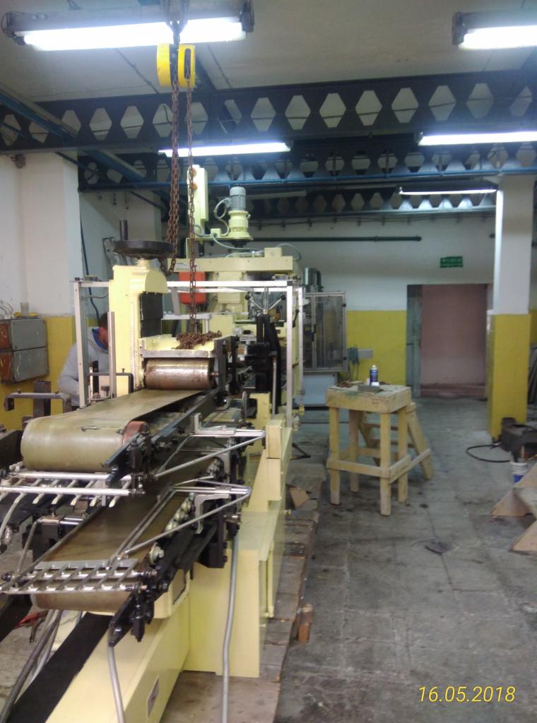 Wnętrze hali z żółtą maszyną produkcyjną z taśmociągiem, podwieszonym łańcuchem i pracownikiem w tle, widok z niskiego kąta.