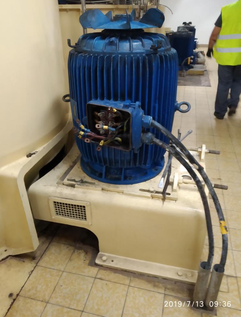 Duży, niebieski silnik elektryczny z otwartą skrzynką przyłączeniową, widoczne kable i narzędzia, w tle osoba w kamizelce odblaskowej.