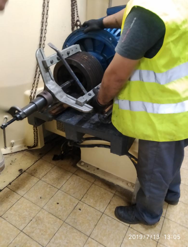 Pracownik w kamizelce odblaskowej demontuje piastę z dużej, niebieskiej maszyny przemysłowej za pomocą specjalistycznego ściągacza; podłoga wyłożona płytkami ceramicznymi w tle.