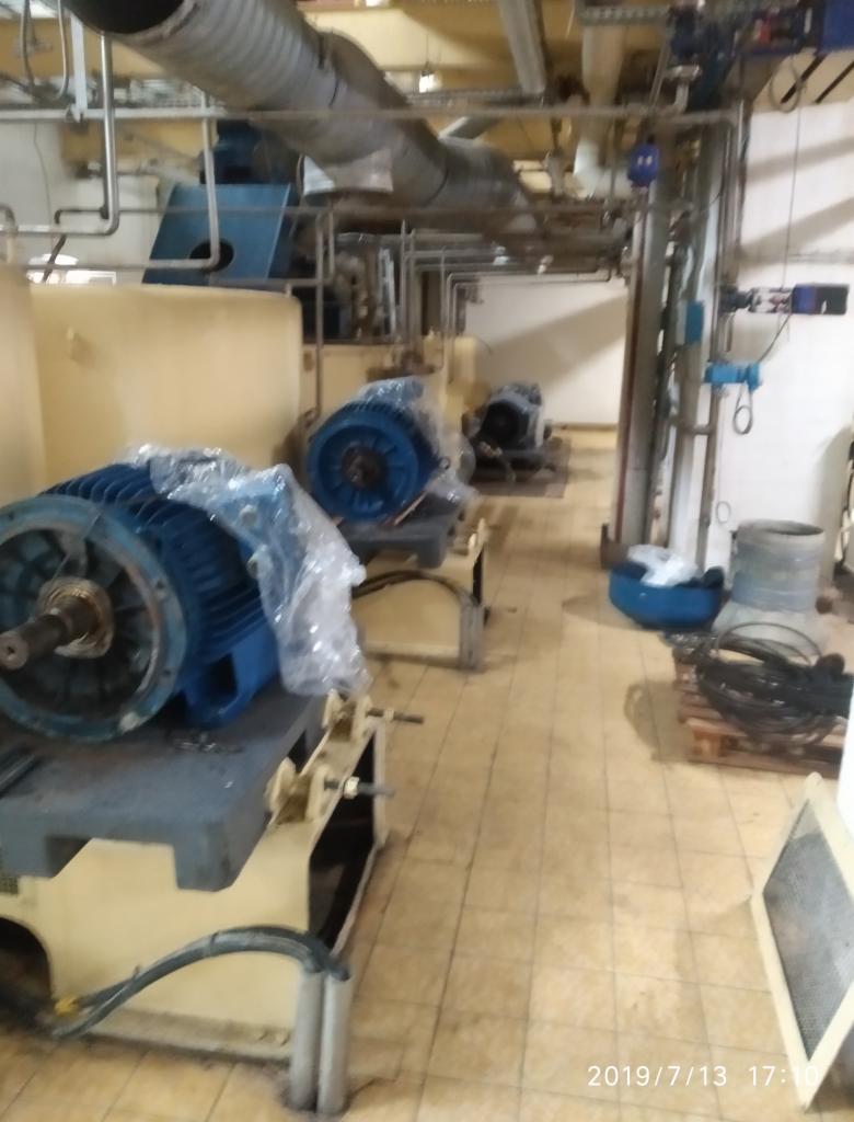 Wnętrze hali produkcyjnej z dużymi, niebieskimi silnikami elektrycznymi, częściowo owiniętymi folią, stojącymi na metalowych podstawach; widoczne rury i instalacje pod sufitem, oraz płytki...