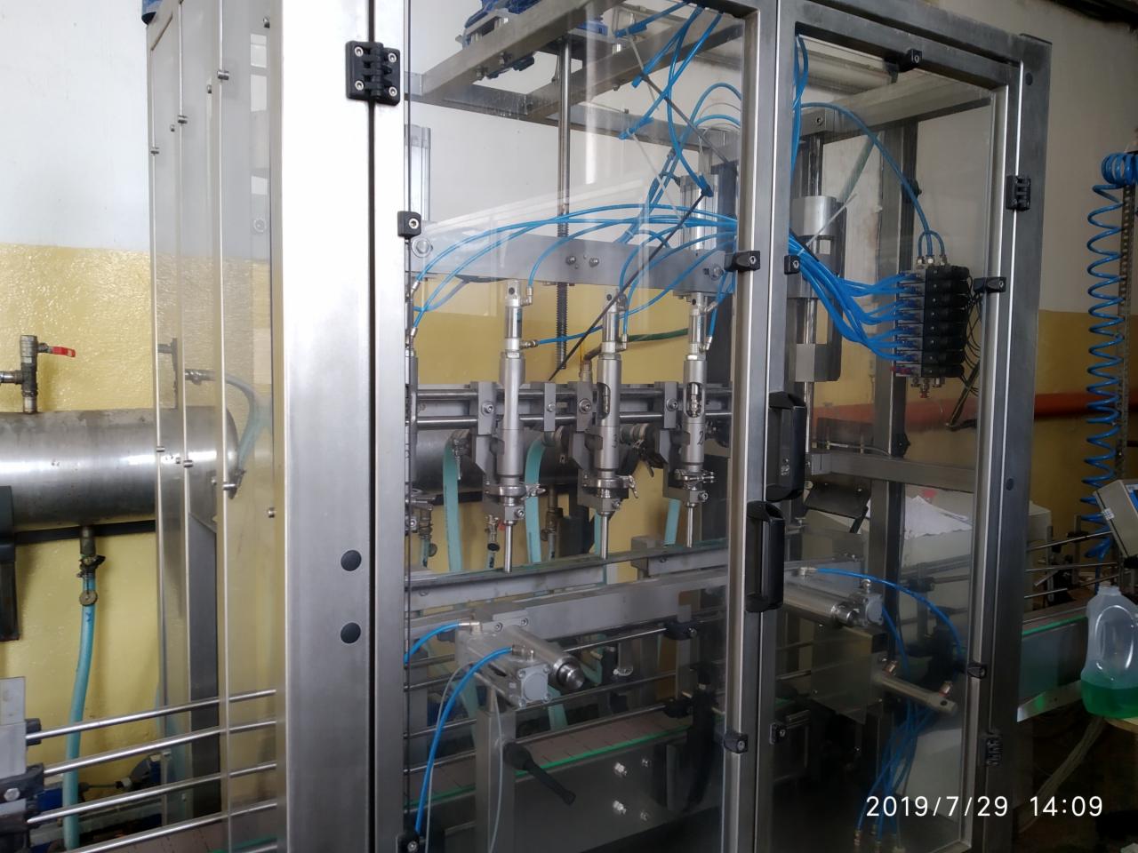 Automatyczna linia rozlewnicza z czterema głowicami, widoczne niebieskie przewody pneumatyczne i metalowa konstrukcja obudowy.