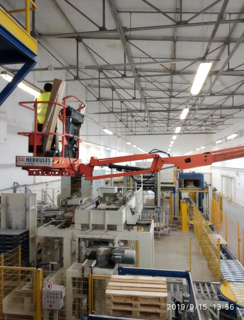 Pracownik w pomarańczowym podnośniku koszowym z logo AA Herkules, wykonujący prace na wysokości w hali produkcyjnej z dużą maszyną przemysłową i paletami w tle.