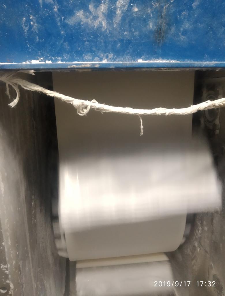 Biały materiał przesuwający się przez maszynę z niebieską obudową, widoczne ślady użytkowania i sznurek w górnej części.