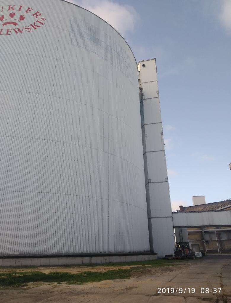 Zewnętrzny widok silosu przemysłowego z blachy falistej z czerwonym napisem 'CUKIER LEWSKI', widoczna drabina i chodnik komunikacyjny, ujęcie z dołu.