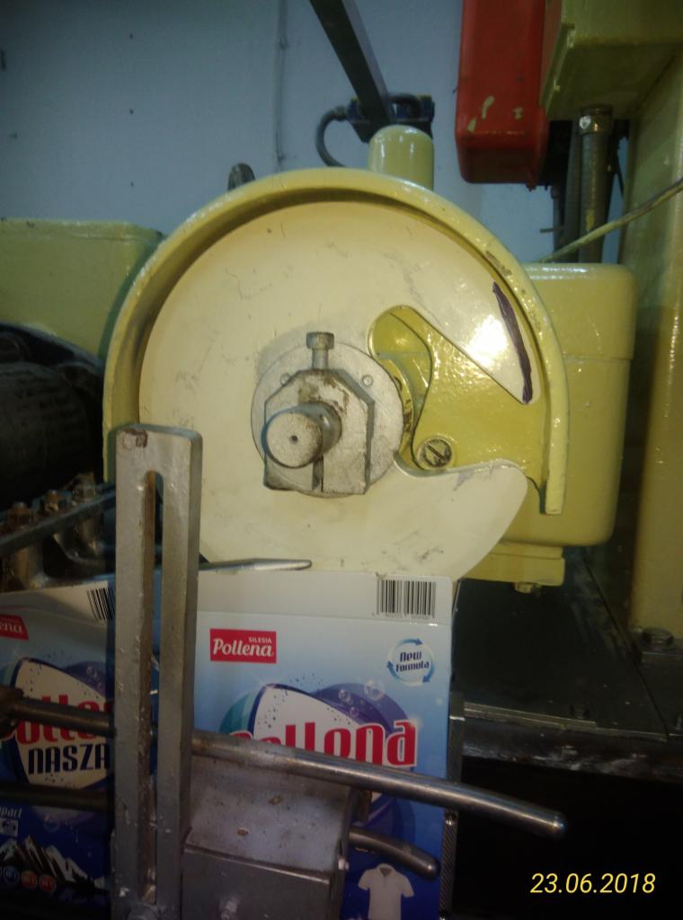 Fragment maszyny przemysłowej z żółtą obudową tarczy, z widocznym mechanizmem mocującym i pudełkiem proszku do prania w tle, data 23.06.2018 widoczna w prawym dolnym rogu.