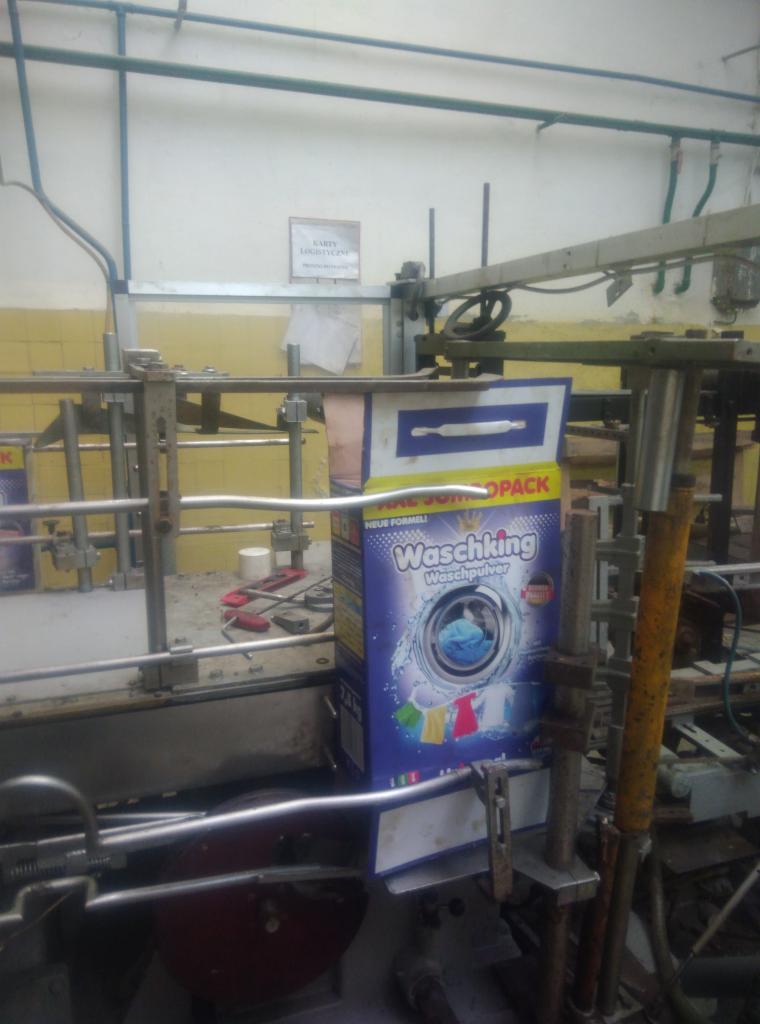 Pudełko proszku do prania 'Waschking' na linii produkcyjnej maszyny pakującej, widoczne elementy mechaniczne maszyny i fragment tabliczki informacyjnej na ścianie w tle.