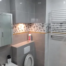 Szara łazienka z geometrycznym wzorem płytek, podświetleniem LED nad toaletą, białym grzejnikiem drabinkowym i otwartą deską sedesową, z płynem do naczyń Ludwik na półce.