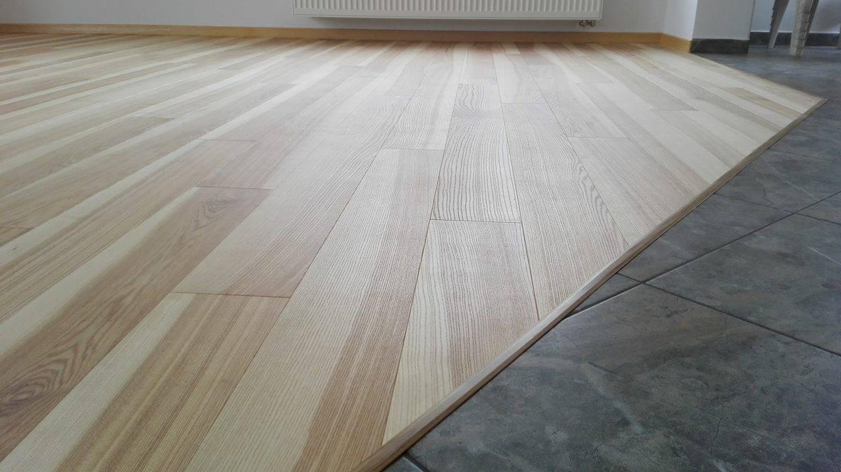 Jasne, drewniane deski podłogowe ułożone wzdłuż ściany, sąsiadujące z szarą, kwadratową podłogą z płytek ceramicznych. Widoczna listwa przypodłogowa i fragment grzejnika na ścianie.