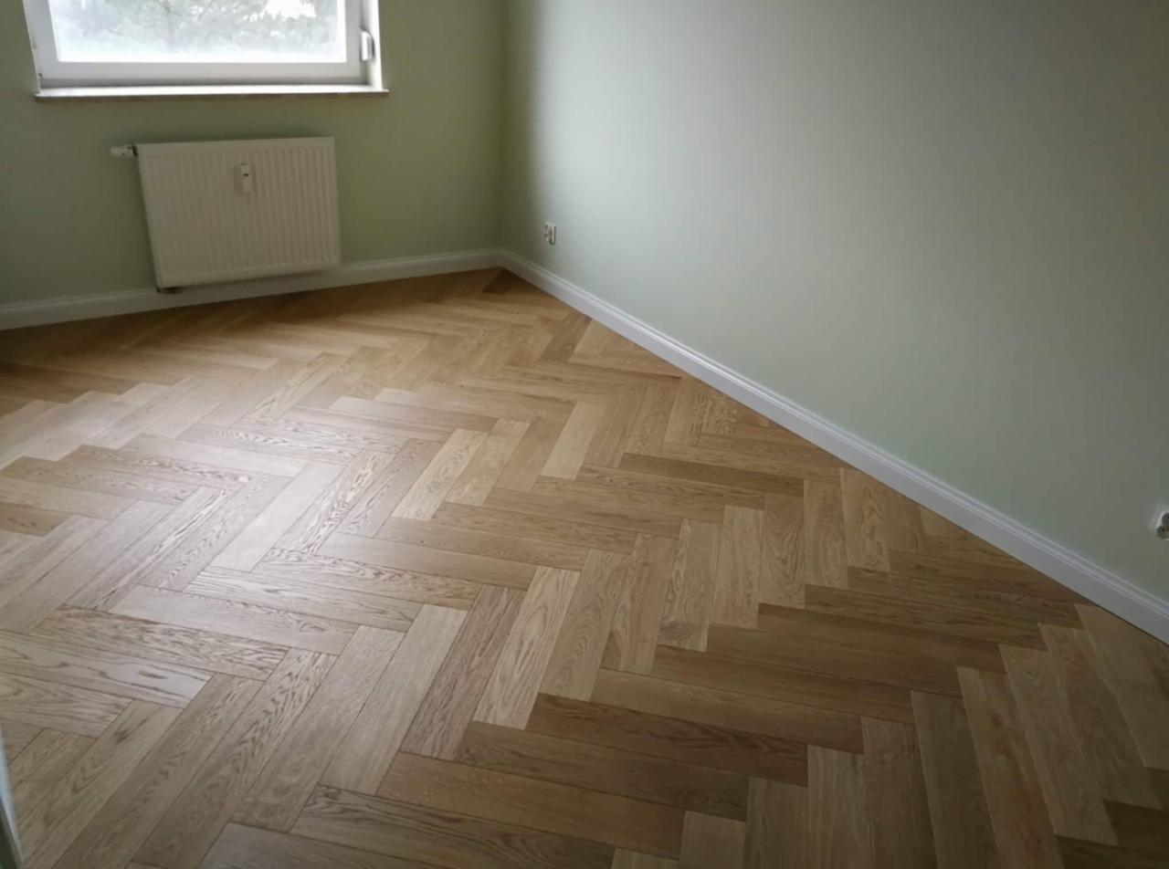 Jasny parkiet ułożony we wzór jodełki francuskiej w pokoju z zielonymi ścianami i białymi listwami przypodłogowymi, widoczny grzejnik pod oknem.