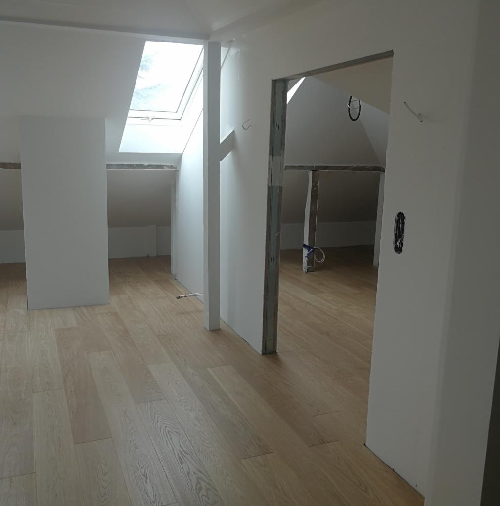 Jasne panele podłogowe ułożone w nowym, pustym pomieszczeniu na poddaszu z oknem dachowym; widoczne niedokończone wykończenia ścian i futryny drzwi.