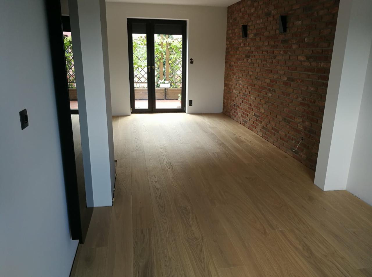 Jasna, dębowa podłoga ułożona w pustym pokoju z ceglaną ścianą i wyjściem na balkon.