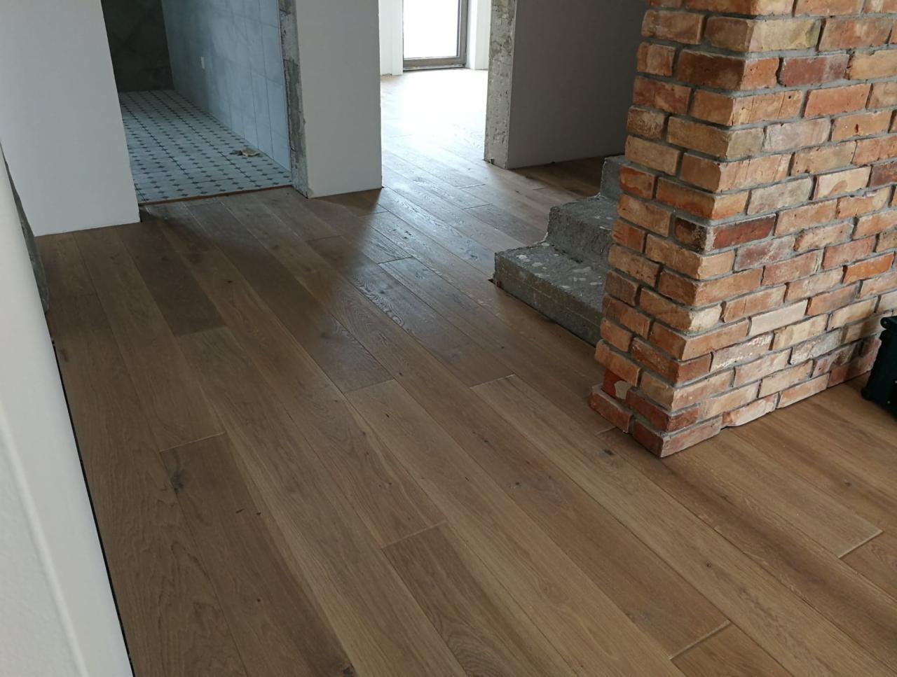 Ukończona podłoga z desek w odcieniach brązu, widoczny fragment ceglanej ściany i surowych betonowych schodów w tle, wnętrze w trakcie wykończenia.