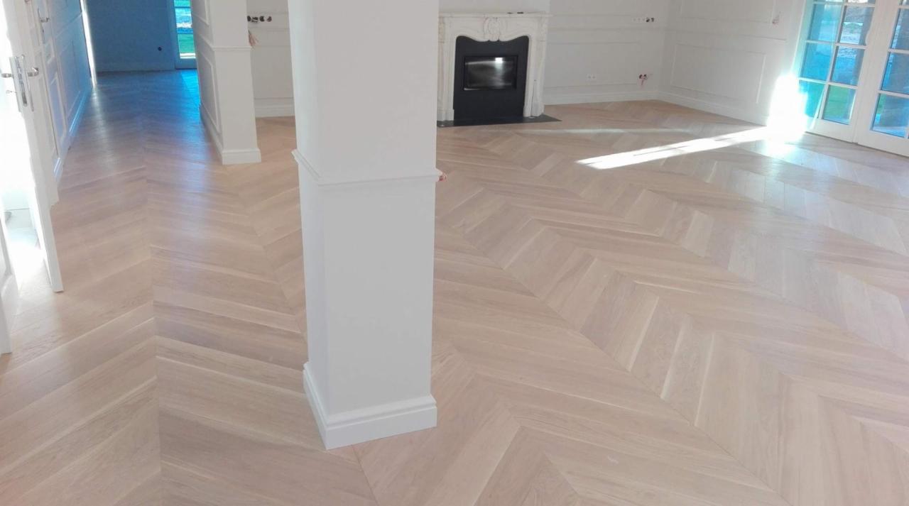 Jasna, drewniana podłoga ułożona w jodełkę francuską w przestronnym, jasnym pomieszczeniu z białymi ścianami i kolumną. W tle widoczny kominek i drzwi balkonowe.