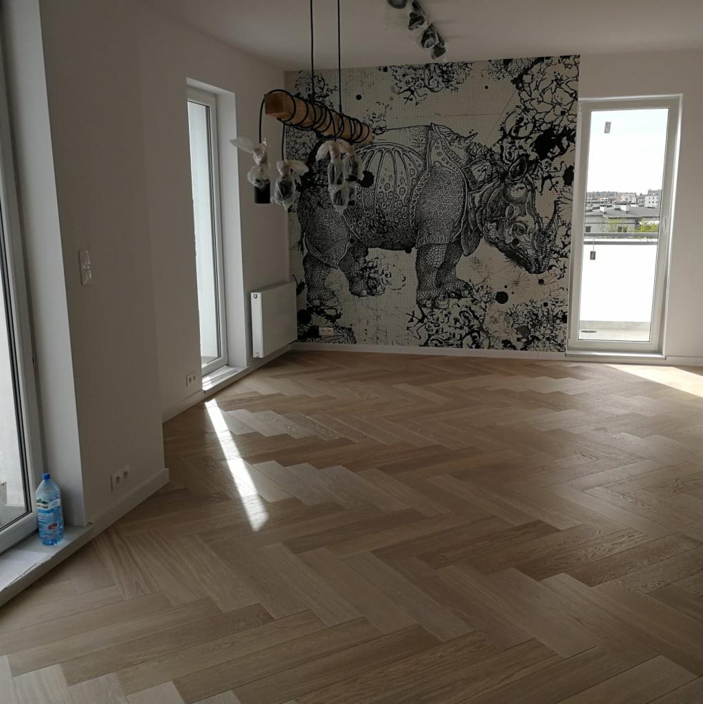 Jasna, drewniana podłoga ułożona w jodełkę francuską w przestronnym pokoju z dekoracyjną tapetą z motywem nosorożca. Światło słoneczne wpada przez okno, tworząc jasną smugę na parkiecie.