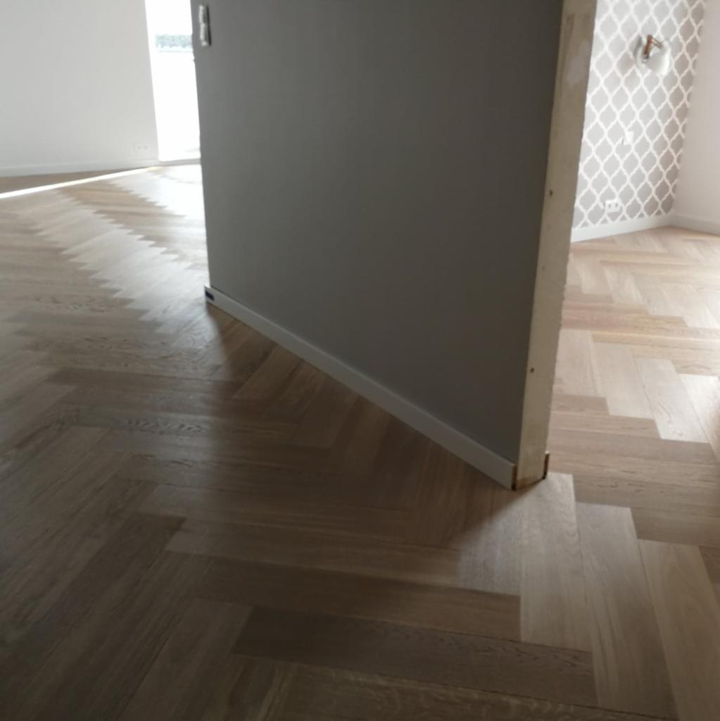 Ułożony parkiet w jodełkę w jasnym odcieniu drewna, widoczny fragment ściany z szarą farbą i białą listwą przypodłogową, oraz fragment ściany z tapetą w geometryczny wzór.