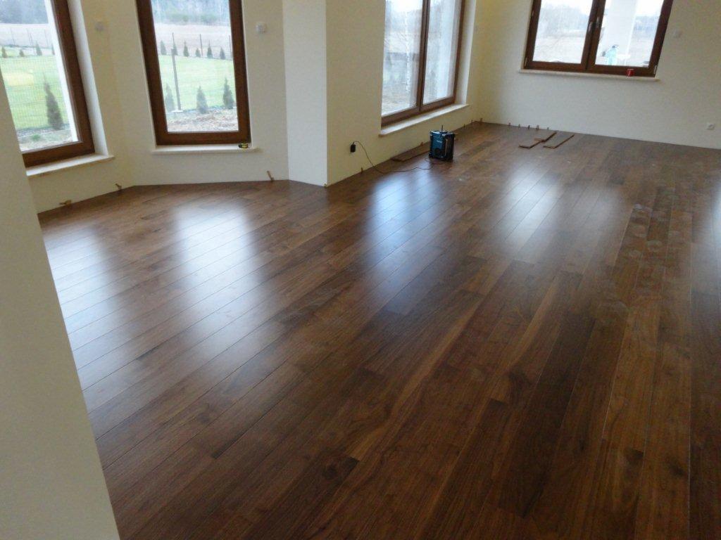 Błyszcząca, ciemnobrązowa drewniana podłoga ułożona w przestronnym pokoju z trzema oknami w drewnianych ramach; widoczny fragment ściany w kolorze kremowym oraz niewielki niebieski sprzęt...