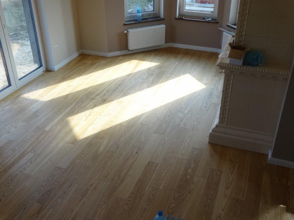 Jasne panele podłogowe ułożone w nowym pokoju z białymi listwami przypodłogowymi, widoczne promienie słoneczne wpadające przez okno i niedokończony piec kaflowy w rogu.