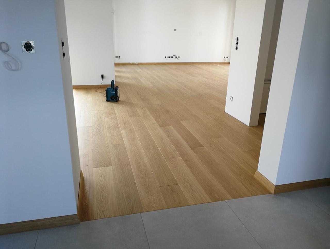 Jasna, dębowa podłoga z desek ułożona w nowym, pustym pomieszczeniu z białymi ścianami i widocznym narzędziem do pomiaru wilgotności powietrza.