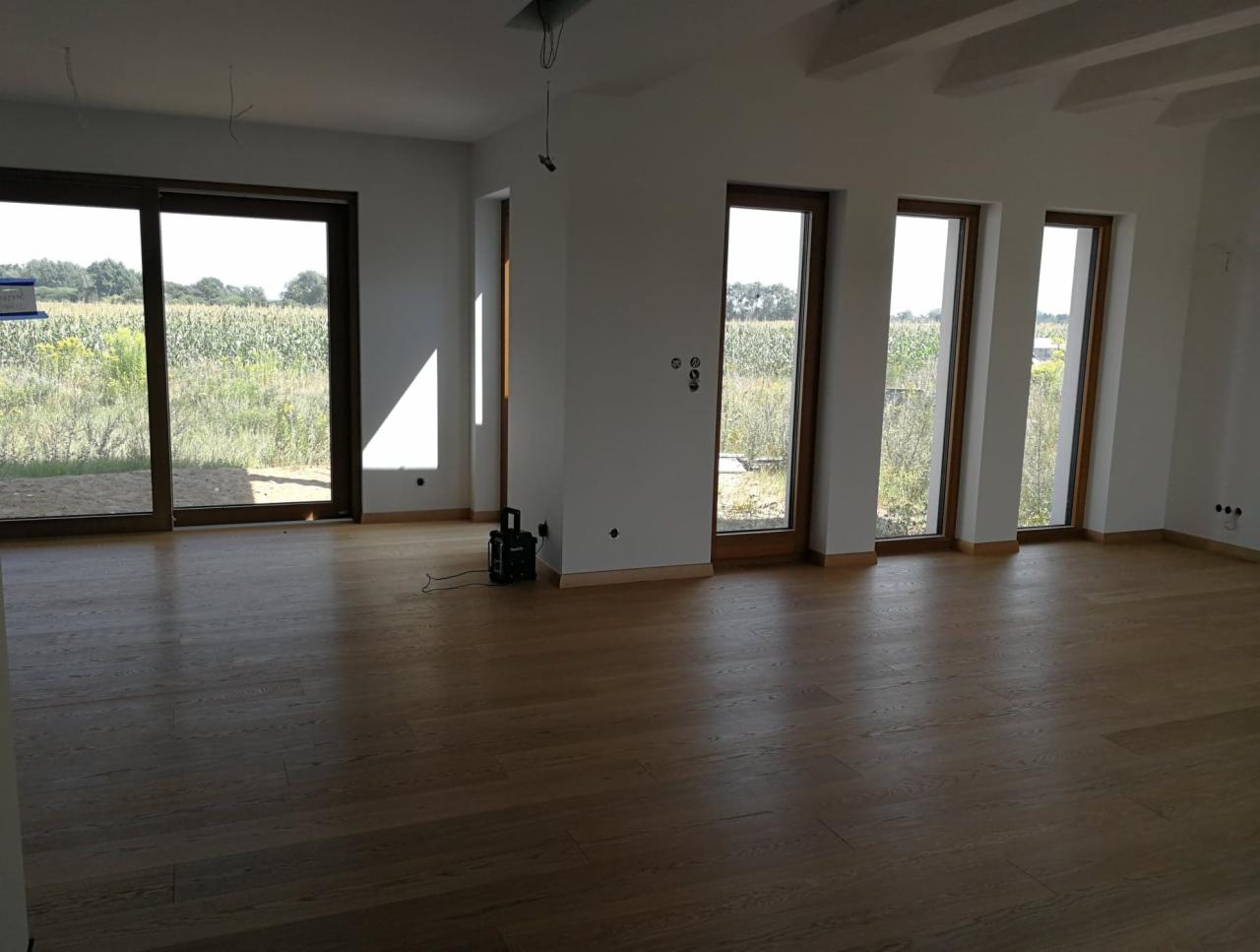 Jasna dębowa podłoga w przestronnym, nowym wnętrzu z dużymi oknami wychodzącymi na pole; na podłodze stoi czarny odkurzacz.