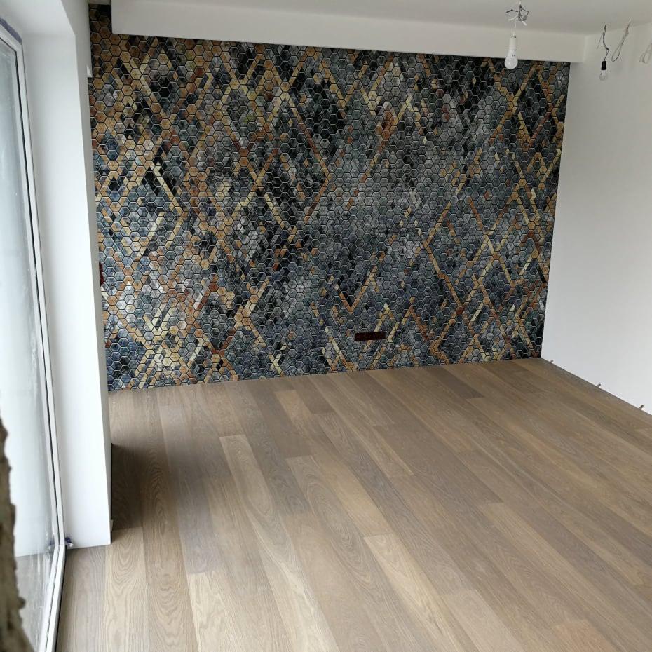 Jasne panele podłogowe w nowym wnętrzu z mozaiką heksagonalną na ścianie w odcieniach szarości, brązu i złota, tworzącą geometryczny wzór w kształcie rombów.