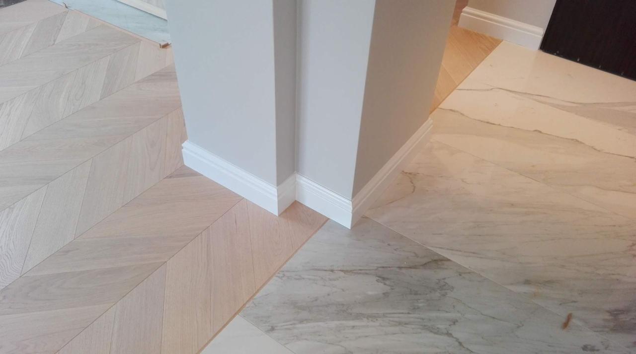Połączenie jasnego parkietu ułożonego we wzór jodełki z marmurową posadzką przy narożniku ściany z białą listwą przypodłogową.