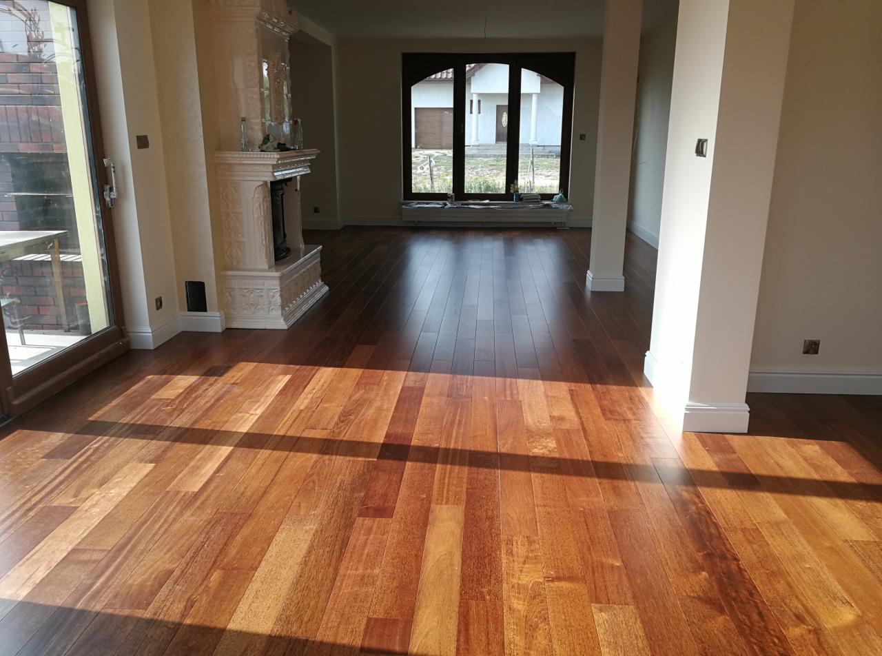 Salon z nową, błyszczącą, drewnianą podłogą z egzotycznego drewna, oświetloną promieniami słońca wpadającymi przez okno. W tle widoczny kominek kaflowy i okno z widokiem na dom.