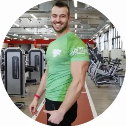 Uśmiechnięty mężczyzna w zielonej koszulce z logo w siłowni, na tle sprzęt do ćwiczeń i bieżnia z czerwoną nawierzchnią.