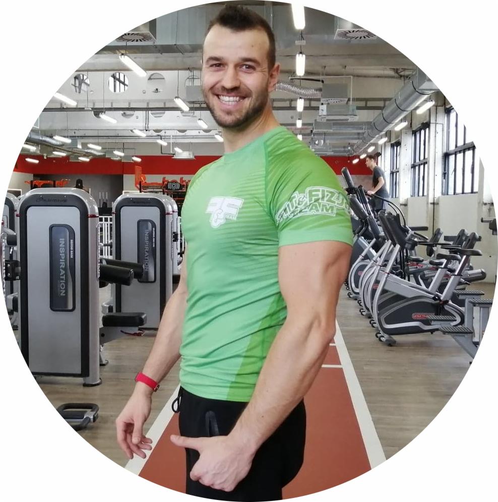Uśmiechnięty mężczyzna w zielonej koszulce z logo w siłowni, na tle sprzęt do ćwiczeń i bieżnia z czerwoną nawierzchnią.