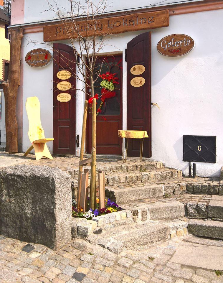 Wejście do galerii z drewnianymi drzwiami, ozdobione kwiatami i tabliczkami informacyjnymi, z kamiennymi schodami i młodym drzewkiem w osłonie.
