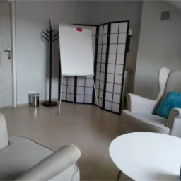 Jasne wnętrze gabinetu z dwoma fotelami, białym stolikiem, flipchartem, parawanem i lampą stojącą. Na stoliku pudełko chusteczek.