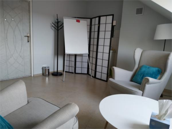 Jasne wnętrze gabinetu z dwoma fotelami, białym stolikiem, flipchartem, parawanem i lampą stojącą. Na stoliku pudełko chusteczek.