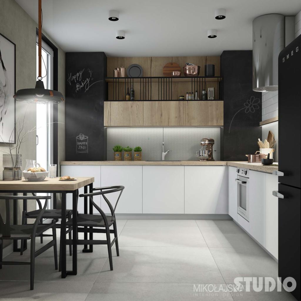 Nowoczesna kuchnia z czarną tablicą kredową, białymi szafkami i drewnianymi akcentami, uzupełniona o czarne krzesła i stół z drewnianym blatem, oświetlona wiszącą lampą w stylu industrialnym.