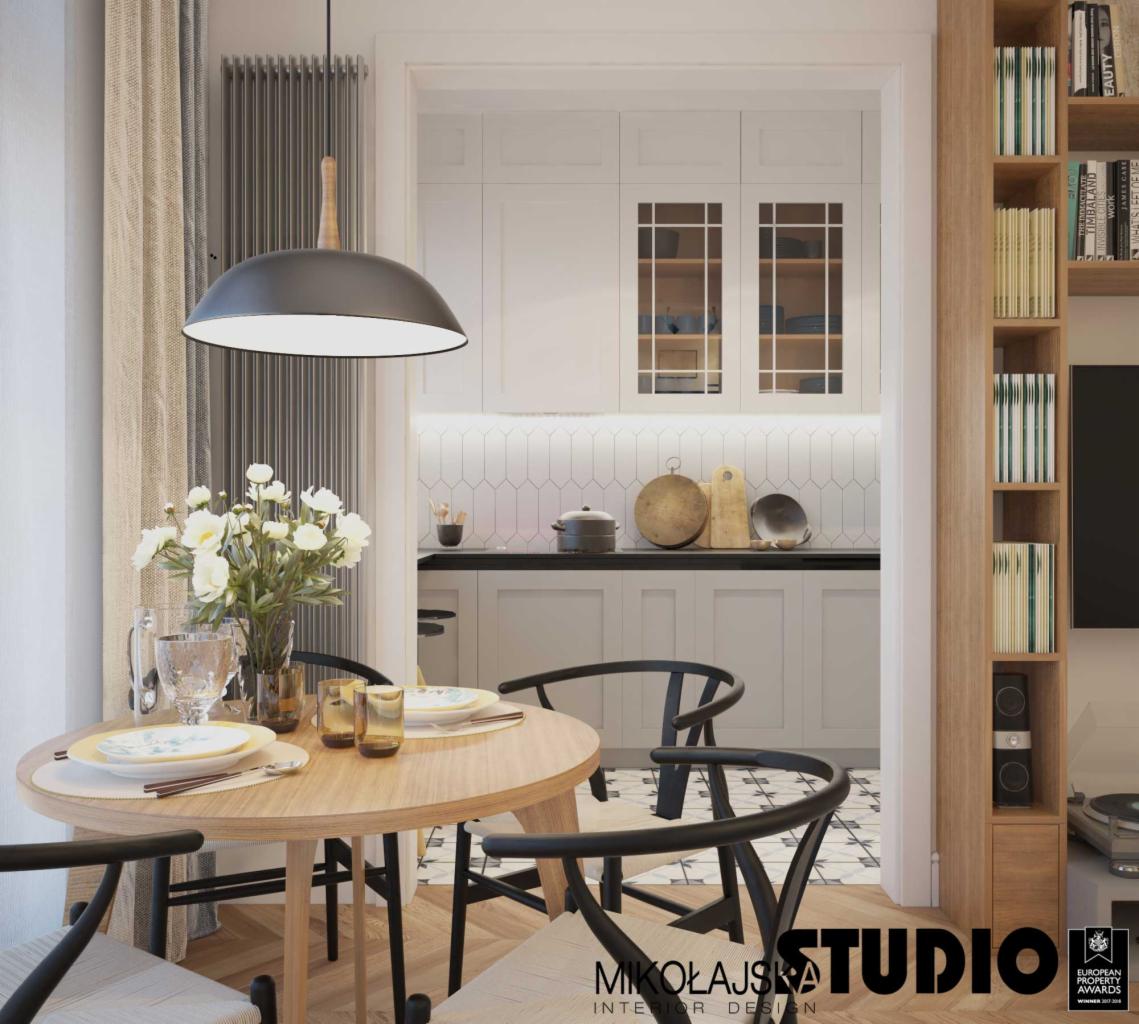 Widok na kuchnię z jadalni: okrągły drewniany stół z nakryciem, czarne krzesła typu Y-back, wisząca lampa kopułowa, biała kuchnia z czarnym blatem i mozaiką na ścianie, drewniany regał z książkami.