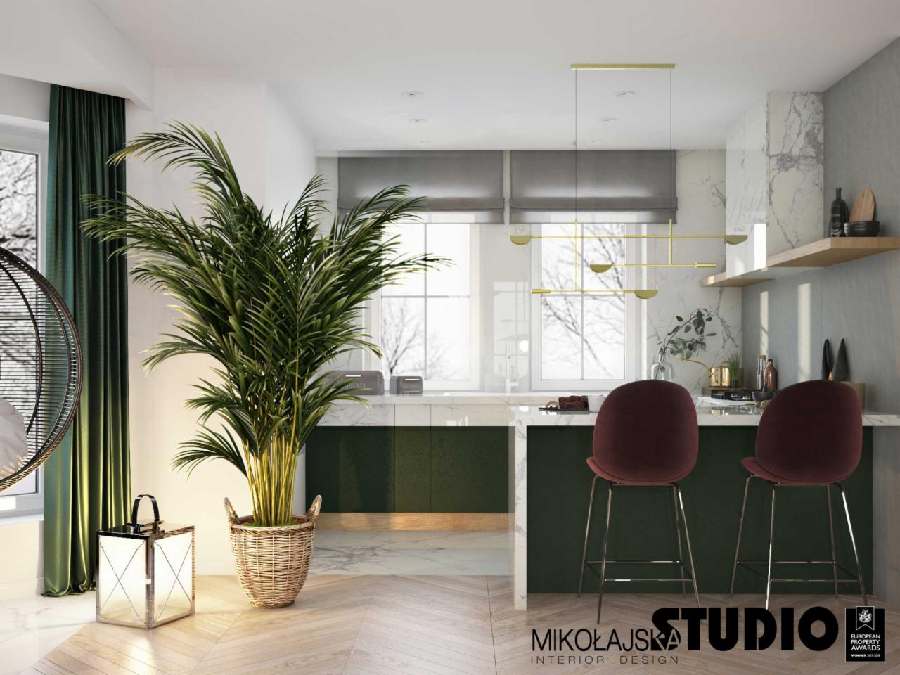 Nowoczesna kuchnia z zielonymi akcentami: wyspa z marmurowym blatem, bordowe hokery, złota lampa wisząca i duża palma w plecionym koszu. Podłoga z jasnego drewna ułożona w jodełkę.
