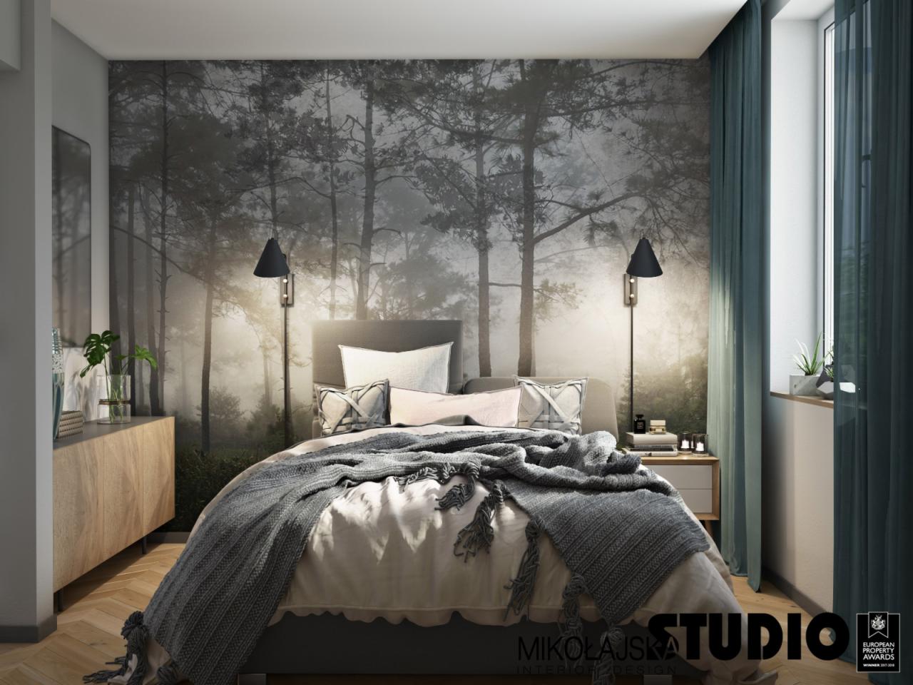 Sypialnia w Krakowie z fototapetą przedstawiającą mglisty las, oświetlona czarnymi kinkietami, z drewnianą komodą i szarą narzutą na łóżku.