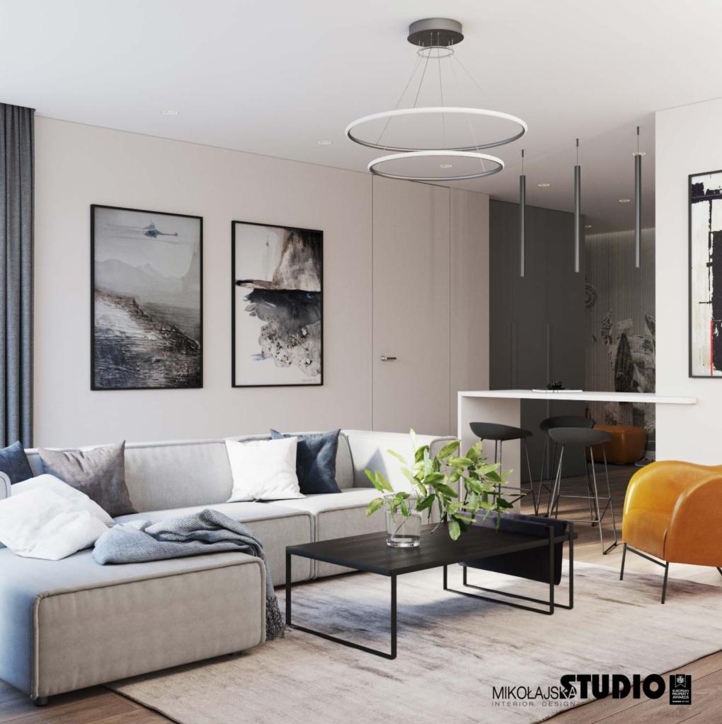 Jasny salon z narożnikiem, stolikiem kawowym z rośliną, okrągłą lampą wiszącą i obrazami na ścianie, utrzymany w minimalistycznym stylu z akcentami szarości i żółci.