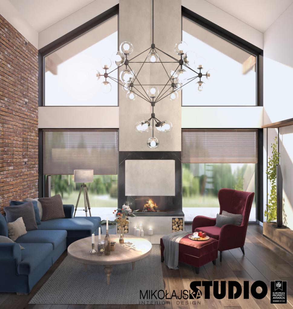 Salon z wysokim sufitem, ceglaną ścianą, kominkiem, nowoczesnym żyrandolem i niebieską sofą narożną oraz bordowym fotelem z podnóżkiem, oświetlony naturalnym światłem wpadającym przez duże okna.