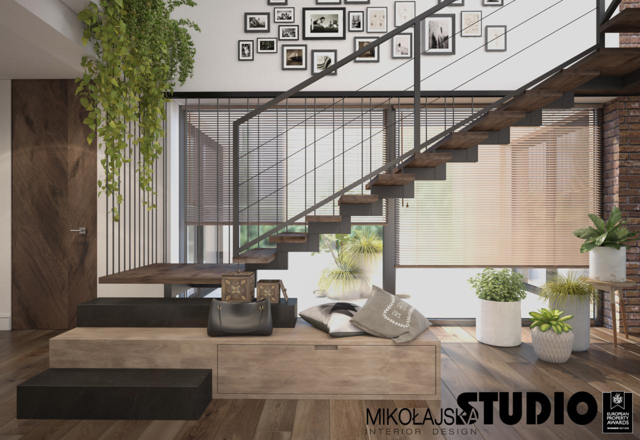 Nowoczesne wnętrze z drewnianymi schodami z metalową balustradą, wiszącymi roślinami, dekoracyjnymi poduszkami i torebką na drewnianej ławce z szufladą, oraz galerią czarno-białych zdjęć na ścianie.