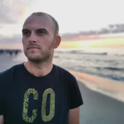 Mężczyzna w ciemnej koszulce z napisem 'CO' na tle plaży o zmierzchu, patrzy w lewo. Widoczne sylwetki ludzi i morze w oddali. Delikatne, ciepłe światło zachodzącego słońca.