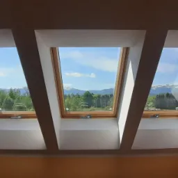 Trzy okna dachowe z drewnianymi ramami, przez które widać błękitne niebo, zielone drzewa i odległe góry.