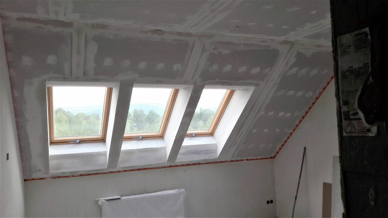 Poddasze w trakcie prac wykończeniowych, widoczne okna dachowe, płyty gipsowo-kartonowe na skosach i suficie, częściowo zaszpachlowane łączenia, zabezpieczone taśmą narożniki.