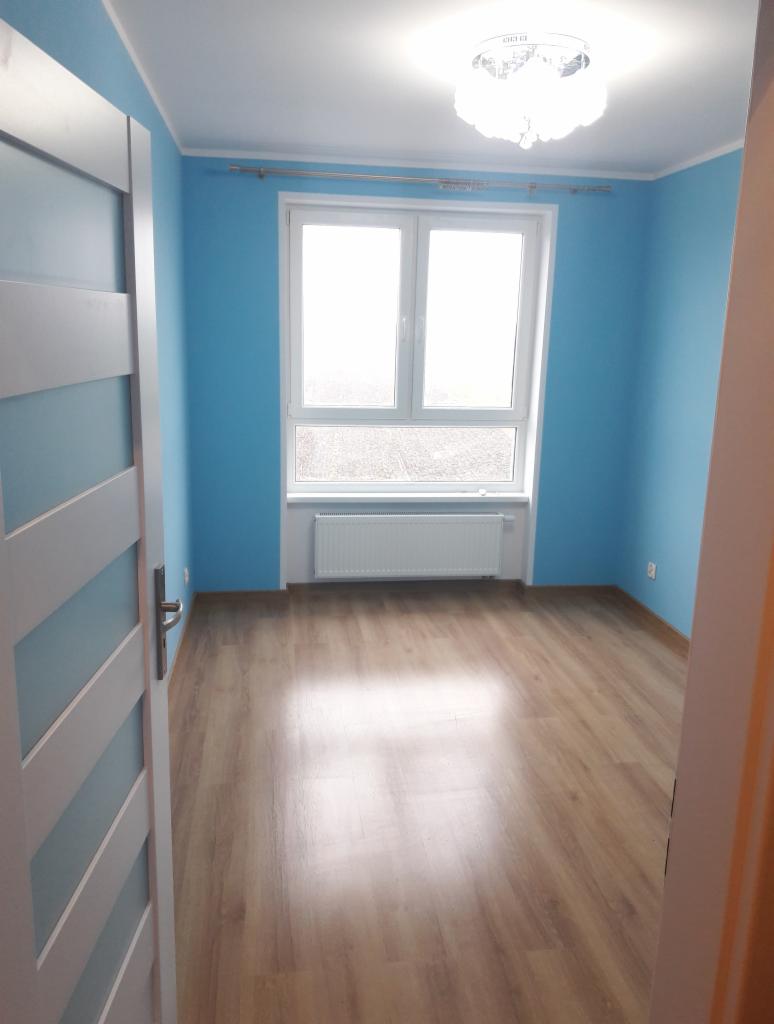 Pusty pokój po odświeżeniu, z niebieskimi ścianami, panelami podłogowymi i białym grzejnikiem pod oknem, widok zza uchylonych białych drzwi z poziomymi panelami.