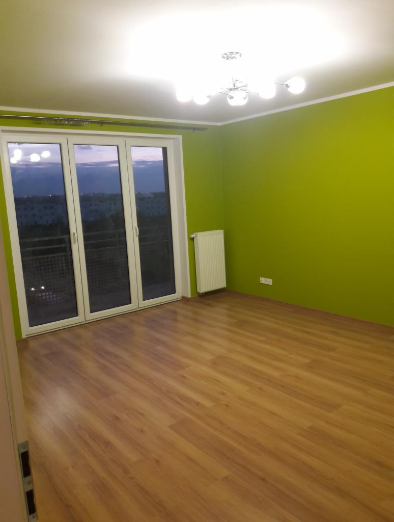 Pusty pokój po odświeżeniu: świeżo pomalowane zielone ściany, nowe panele podłogowe, białe drzwi balkonowe i grzejnik.