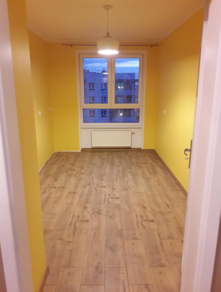 Pomalowany na żółto pokój z drewnianą podłogą, widok na okno z grzejnikiem pod nim i lampą sufitową.