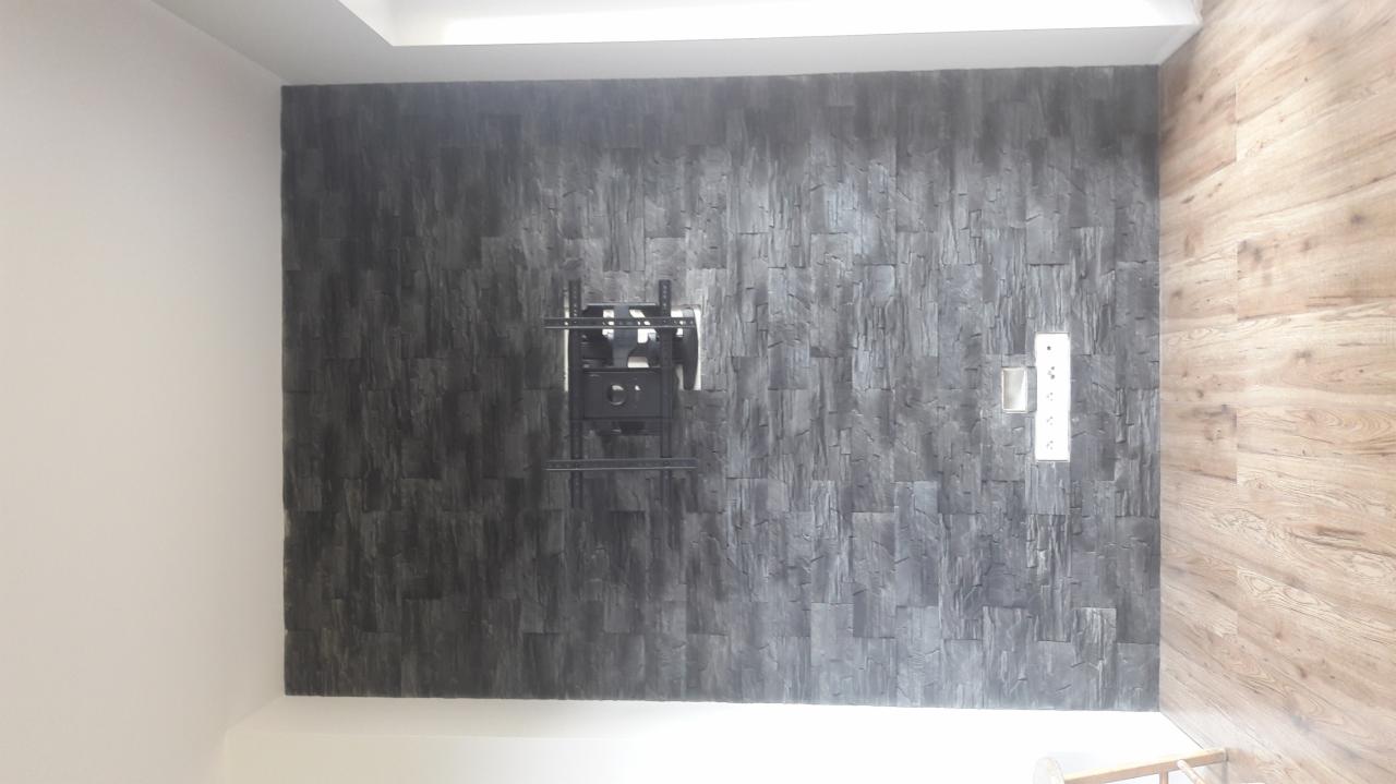 Ściana wykończona szarym kamieniem dekoracyjnym z zamontowanym uchwytem do telewizora i gniazdkiem elektrycznym, sąsiadująca z ścianą wyłożoną panelami drewnopodobnymi.